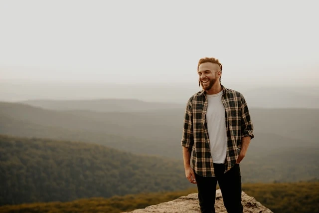 Homme souriant debout sur une crête rocheuse, chaînes de montagnes boisées à l'arrière-plan